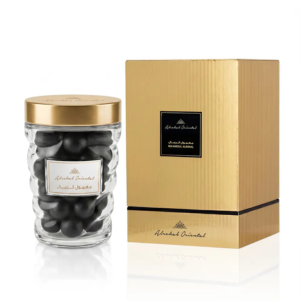 A clear glass jar filled with dark round chocolates, topped with a gold lid, shown next to its metallic gold box packaging on a white background.