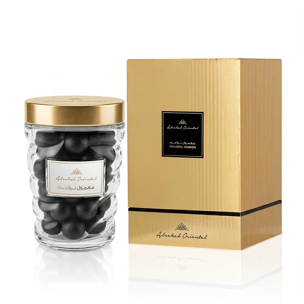 A clear glass jar filled with dark round stones or beads, topped with a metallic gold lid, shown beside a tall gold presentation box with a black label. The brand name 'Abdel Aziz' is visible on both the jar label and the box.