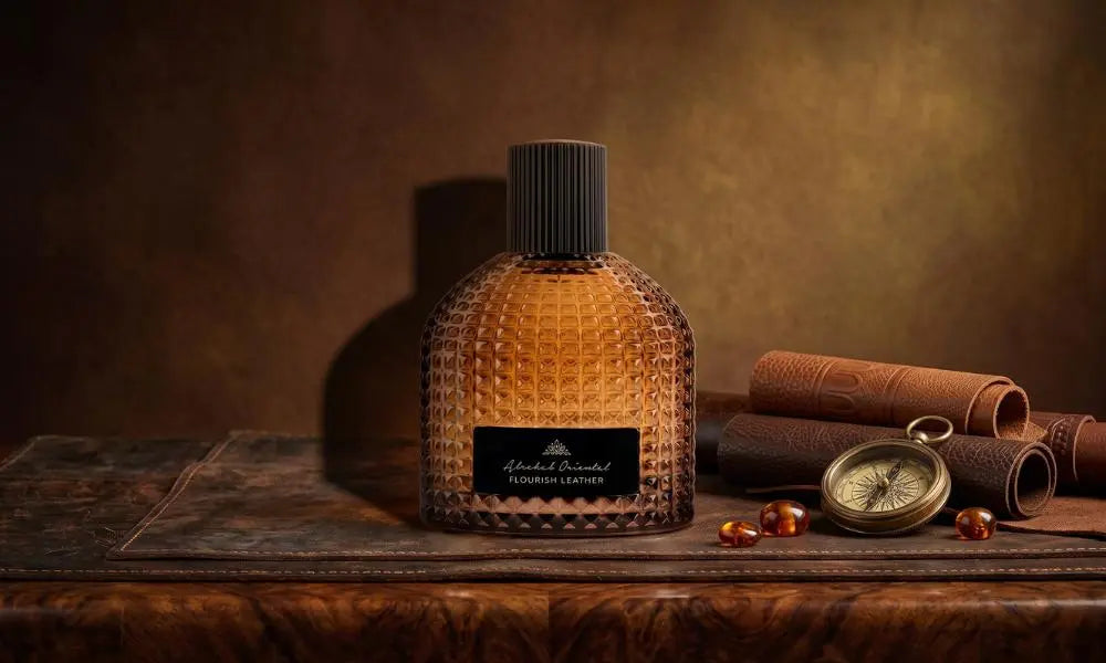 A textured amber perfume bottle, labeled "Flourish Leather," sits on a dark leather mat next to rolled leather, amber beads, and an vintage compass.