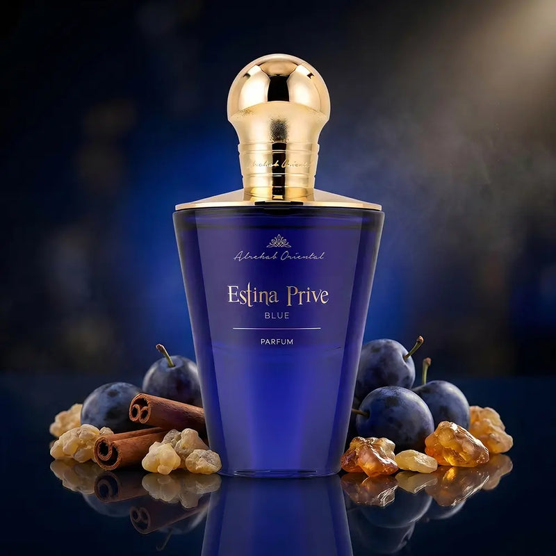 A bottle of Estina Prive Blue Parfum by Molecule Oriented, featuring a deep blue glass body and a metallic gold cap, styled with blue plums, cinnamon sticks, and amber resin on a reflective dark surface.