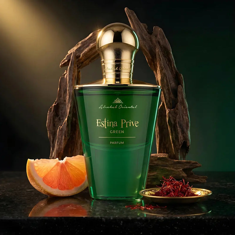 A green glass perfume bottle labeled 'Estina Prive Green' with a shiny gold cap, styled on a dark surface with a grapefruit slice, dried branches, and a small dish of red spice against a dark green background.