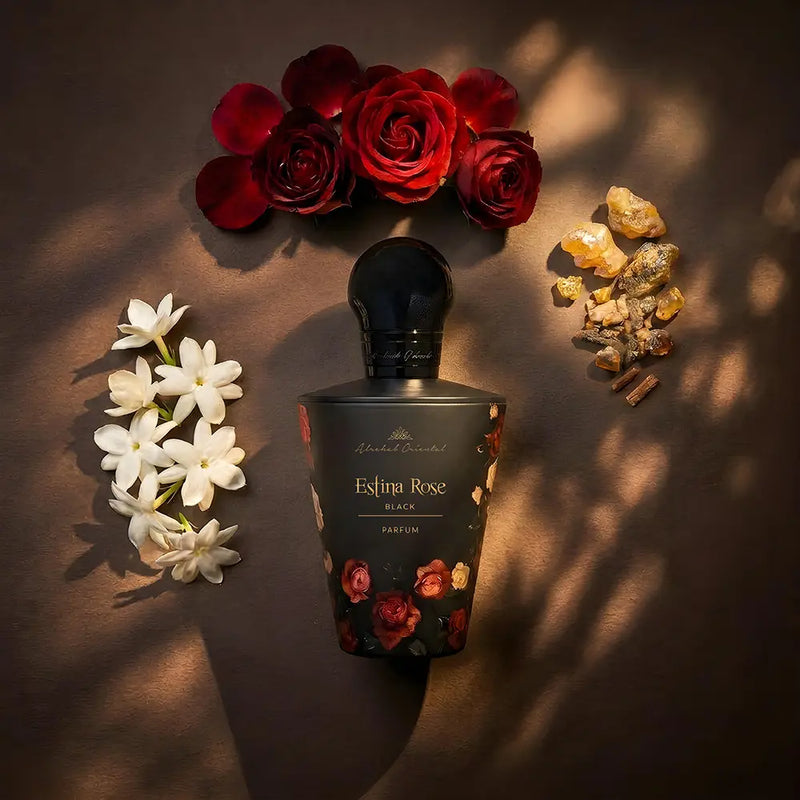 A perfume bottle labeled 'Elixir Rose Black' is centered on a dark brown surface, surrounded by red roses, white jasmine flowers, and small amber/gold botanical pieces. The bottle is matte black with floral artwork near the bottom and a glossy black cap.