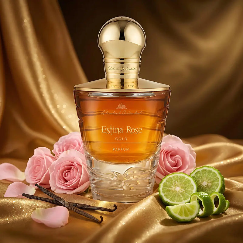 A perfume bottle labeled 'Esfina Rose GOLD' with amber liquid and a shiny gold cap, styled on draped gold fabric with pink roses, lime slices, and vanilla beans.