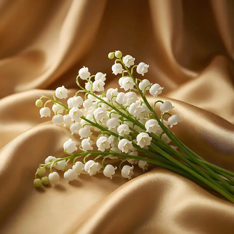 A bouquet of white lily of the valley flowers with green stems resting on draped champagne-colored satin fabric.