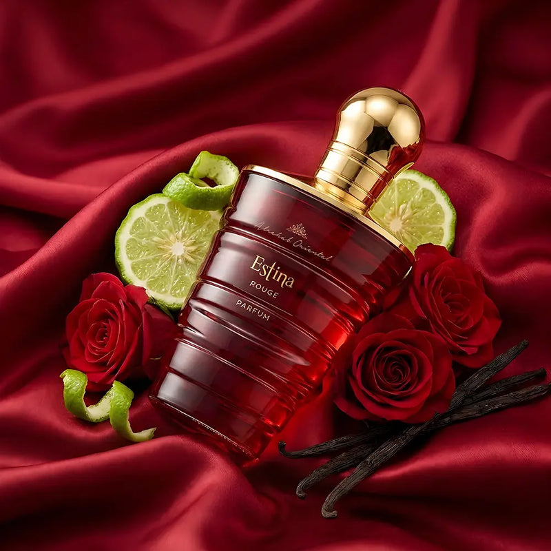 A bottle of Estina Rouge perfume by Chantilly Paris, featuring a dark red translucent glass body and a shiny gold cap, styled with red roses, lime slices, vanilla beans, and green zest on a draped red satin background.