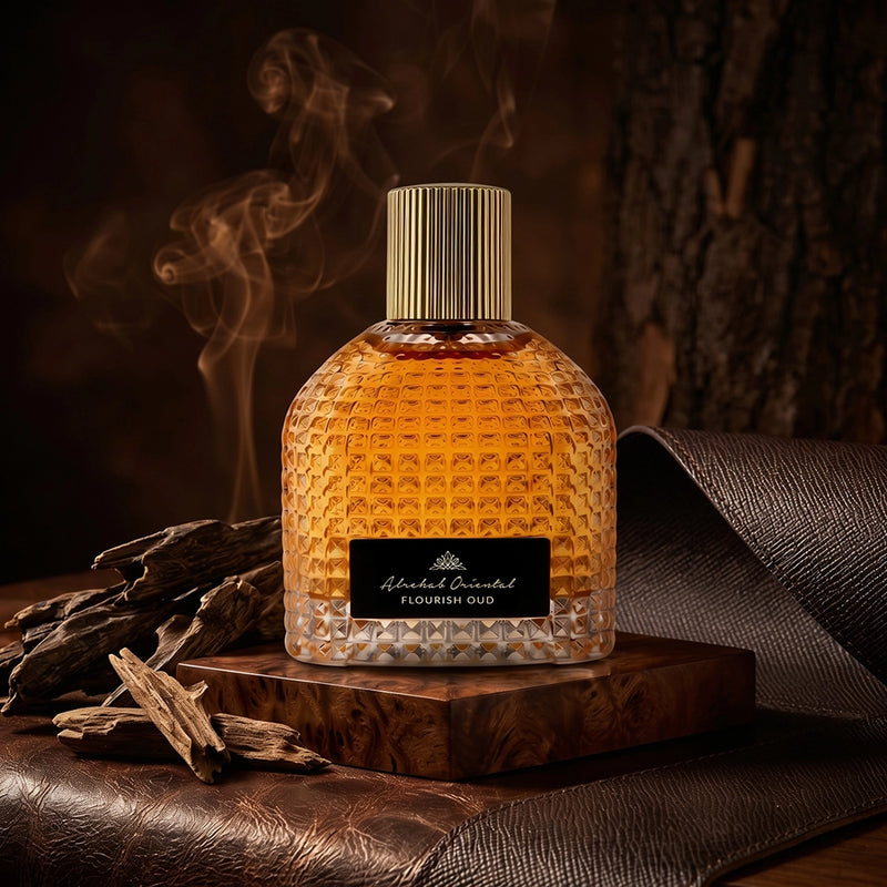 A textured glass perfume bottle filled with amber liquid, featuring a ribbed gold cap and a black label reading 'Aarav Sharma' and 'Flourish Oud'. The bottle is styled on a wooden block with dried wood pieces and a rolled leather sheet against a dark, atmospheric background with smoke.