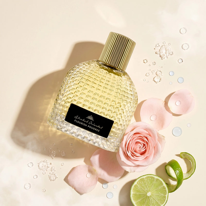 A textured glass perfume bottle with a gold cap and a black label reading 'Aldamare & Co. FLOURISHING FLOWERS', styled with pink rose petals, a lime slice, lime peel, and water droplets on a warm cream background.