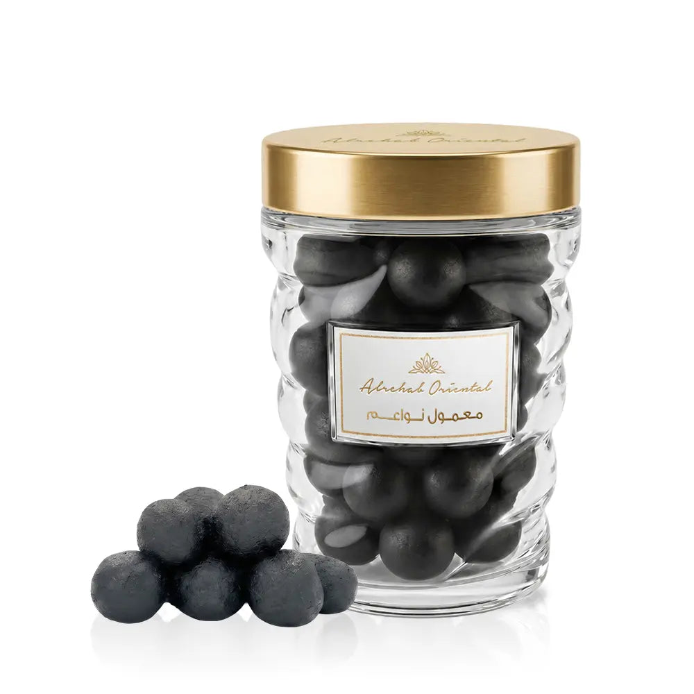 A clear glass jar with a shiny gold lid, filled with dark charcoal-colored round candies, with a few candies spilled in front on a white background. The label reads 'Abrahed Oriental' in English and Arabic.
