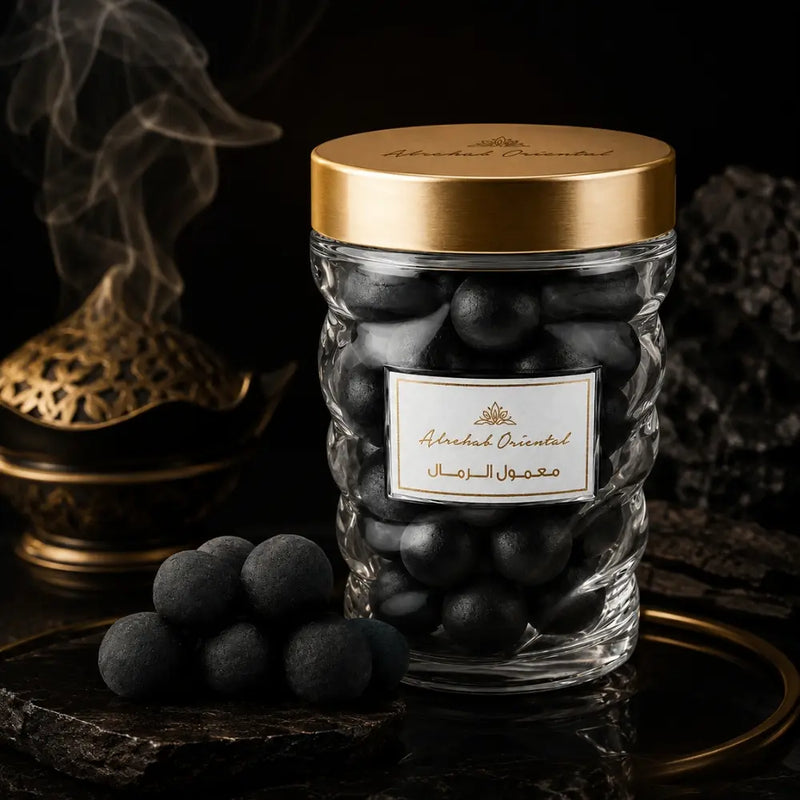 A clear glass jar filled with Maamoul, topped with a metallic gold lid The jar sits on a dark surface with a few matching dark spheres in front, against a moody black background with a brass incense burner and smoke visible.