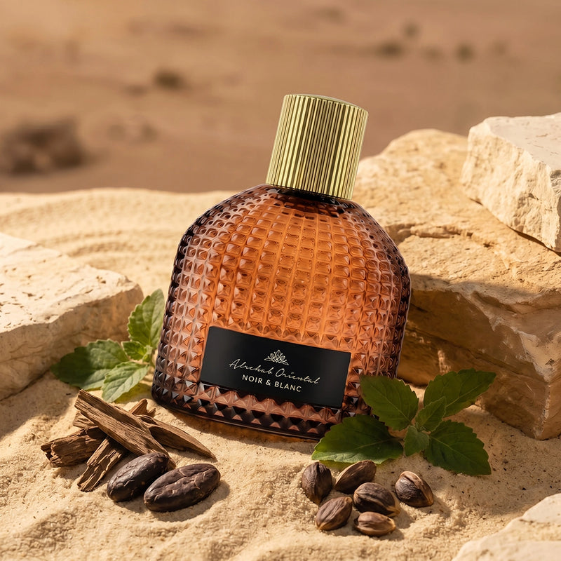 A textured amber glass perfume bottle with a ribbed gold cap and a black label reading 'Aldamand Bruchel NOIR & BLANC', styled on sand with rocks, cinnamon sticks, and green leaves.