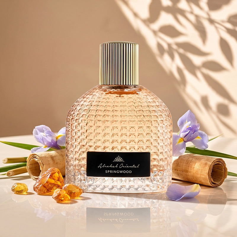 A textured glass perfume bottle with a gold ribbed cap and a black label reading 'Albert Oriental SPRINGWOOD', styled on a reflective surface with purple flowers, amber drops, and rolled paper against a warm beige background with leaf shadows.