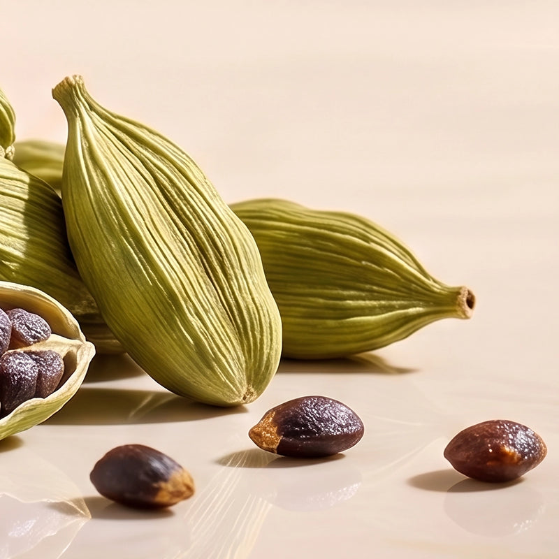 Green cardamom pods with a few dark brown seeds scattered on a light beige surface, with one pod split open showing the seeds inside.