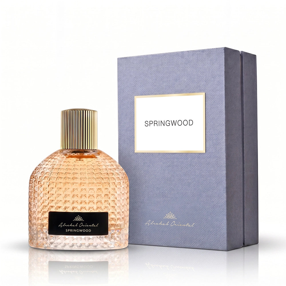 A bottle of Abraham Oriental Springwood perfume with a textured peach-colored glass body and a gold cap, displayed next to its textured gray-blue packaging box with a gold-bordered label.