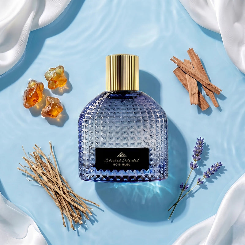 A textured blue glass perfume bottle with a gold cap and a black label reading 'Aromatic Esthetique BOIS BLEU', styled on a light blue water-like surface with white draped fabric, amber stones, cinnamon sticks, and lavender sprigs.